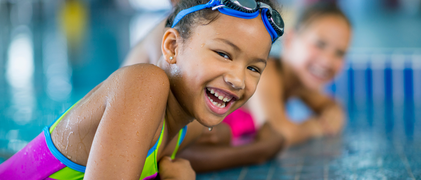 Four kids smiling, standing in front of pool with pool noodles